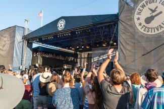 The crowd claps while the stage of the Newport Folk Festival stands in the background on a beautiful day in Newport, RI